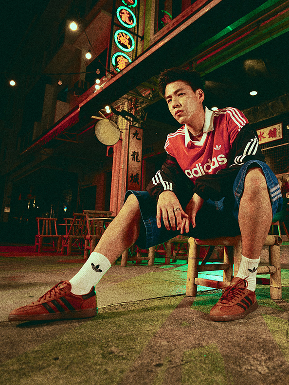 A young man in an Adidas sports jersey sits on a bench in front of a colorful, neon-lit storefront.