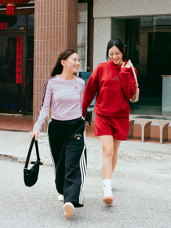 Two young women, one wearing a pink top and the other a red dress, are walking together and smiling as they pass by a brick building with Chinese characters on the sign.