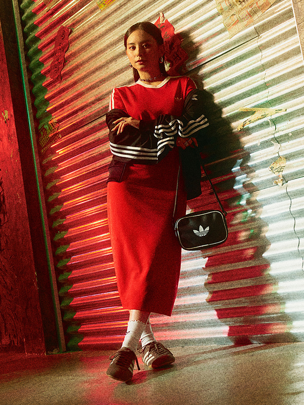 A woman in a red dress stands in front of a colorful, corrugated metal wall, holding a black Adidas bag.