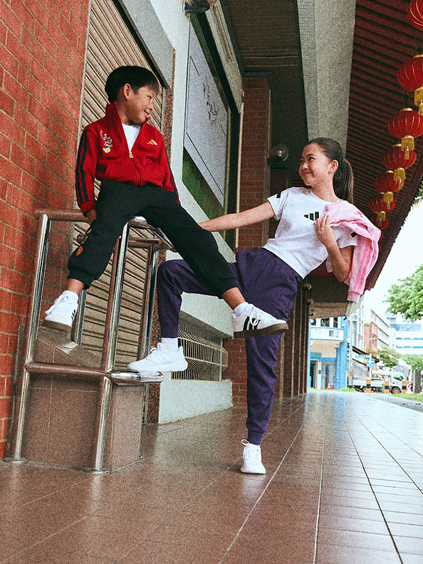 Two young people, a boy and a girl, are sitting on a ledge in what appears to be an outdoor setting with a brick wall and other structures in the background.