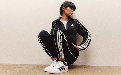 A person wearing a black and white Adidas tracksuit is standing in a plain, light-colored room.
