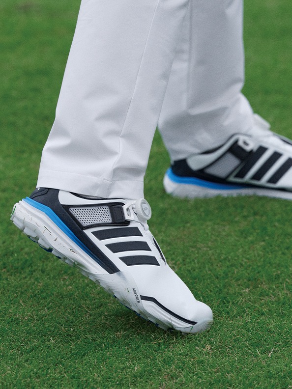 The image shows a person's feet wearing white pants and a pair of black and blue athletic shoes standing on a grassy field.