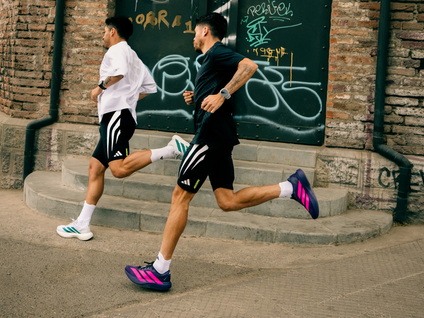Two individuals in athletic attire are running on a sidewalk in front of a graffiti-covered brick wall.