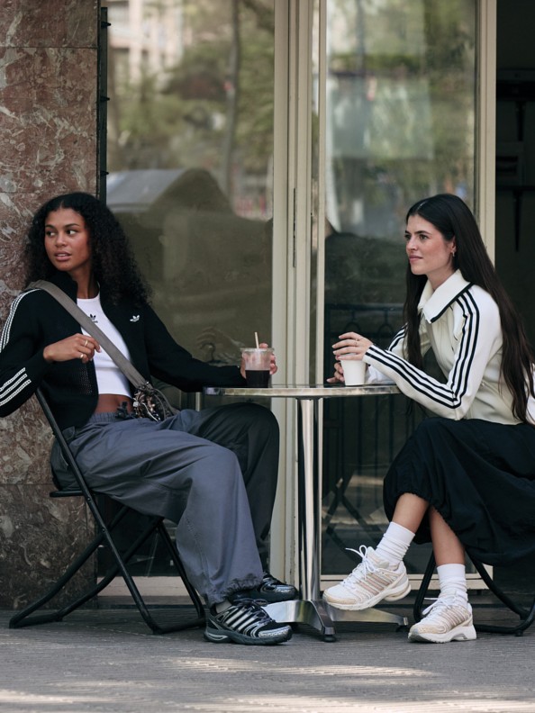 Two young women, one with curly hair and the other with straight hair, are sitting on chairs in front of a large window, enjoying drinks and conversing.