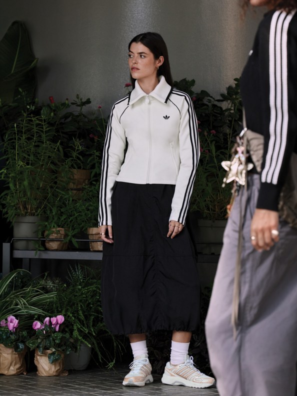 A person wearing a white jacket with black stripes stands in a setting with potted plants and flowers in the background.