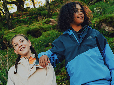 Twee jonge individuen, de ene met gekruld donker haar en de andere met gevlochten haar, staan samen in een weelderige, groene outdooromgeving, omgeven door bomen en vegetatie.