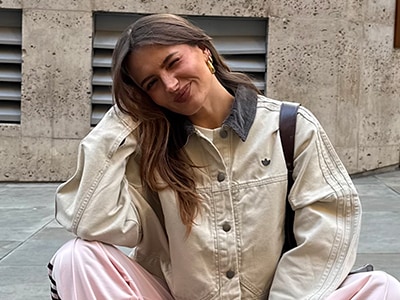 Een jonge vrouw met lang bruin haar zit op een betonnen grond, draagt een beige jas en een roze rok, met een glimlach op haar gezicht.