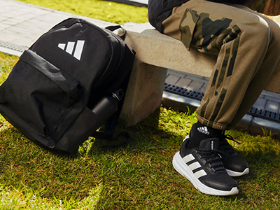 A imagem mostra a zona inferior de uma pessoa a usar sapatilhas adidas e num campo relva, com uma mochila preta e um saco com padrão de camuflado visíveis em primeiro plano.