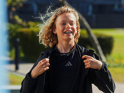 Um jovem com cabelo loiro encaracolado e um sorriso brilhante, usando um casaco preto, destaca-se num ambiente semelhante ao parque com árvores e verde ao fundo.