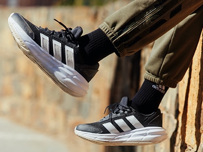 A imagem mostra uma vista aproximada dos pés de uma pessoa a usar sapatilhas adidas pretas e brancas, com o fundo a parecer ser uma superfície ou estrutura de madeira.