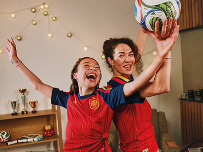 Zwei Frauen in roten und blauen Sporttrikots feiern und heben ihre Arme in Aufregung, mit einem festlichen Hintergrund aus Lichterketten und Trophäen, die hinter ihnen sichtbar sind.