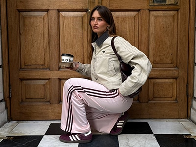 Eine junge Frau in einer weißen Jacke und einer rosa gestreiften Hose steht vor einer Holztür und hält eine Tasse in der Hand.