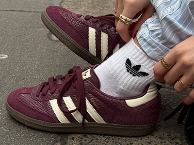Das Bild zeigt adidas Sneaker in Bordeauxrot mit weißen Streifen, die von einer Person getragen werden, die weiße Socken trägt und auf einem Betonbelag steht.