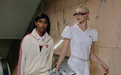 Two individuals, a woman with dark hair and a woman with blonde hair, stand in a dimly lit setting against a textured wall.