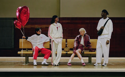 A group of individuals dressed in stylish, coordinated outfits stand on a stage against a plain background, with one person holding a large red heart-shaped balloon.