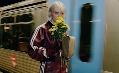 A young woman with blonde hair stands in a doorway, holding a bouquet of yellow flowers and wearing a burgundy jacket.