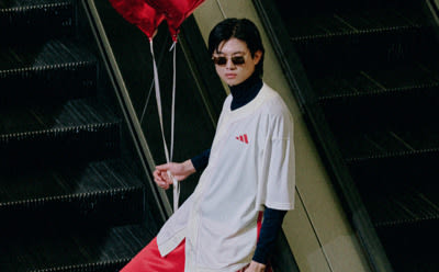 A person wearing a white robe and holding red balloons stands in a dimly lit hallway with a dark background.