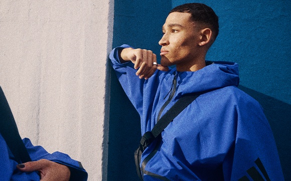 A person wearing a blue hooded jacket stands in front of a blue wall, appearing to be in deep thought or contemplation.