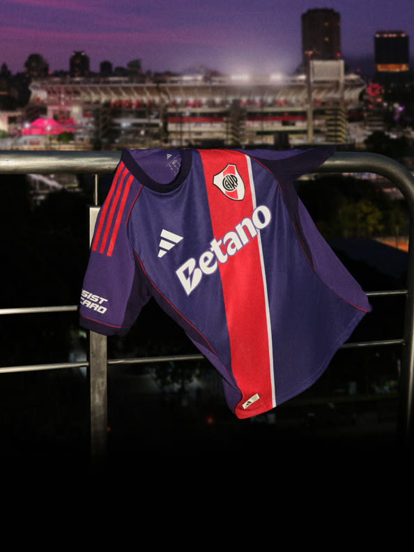 A purple and red soccer jersey hangs on a railing, with a sports stadium visible in the background at night.