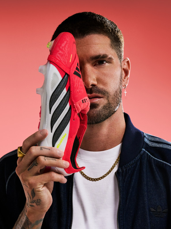 A man with a beard and tattoos holds a red and white sneaker against a vibrant red background.