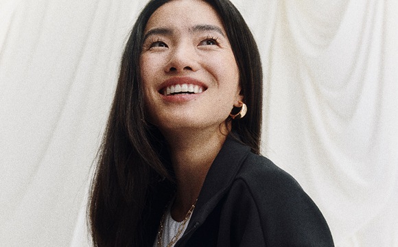 A smiling woman with long dark hair stands in front of a white curtain backdrop, her expression conveying a sense of joy and positivity.