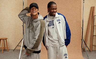 Two young men wearing casual athletic clothing, one in a gray sweatshirt and the other in a white Adidas jersey, standing in what appears to be a locker room or indoor sports facility.