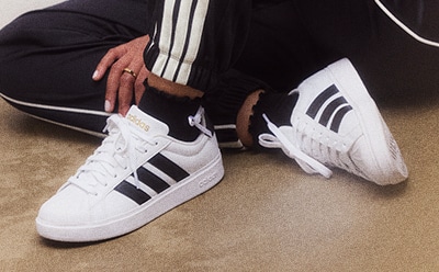 A pair of white and black Adidas sneakers on a carpeted floor, with a person's legs and hands visible in the foreground.