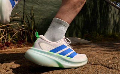 A person's foot wearing a pair of white and blue sneakers with a thick sole stands on a dirt path surrounded by lush greenery.