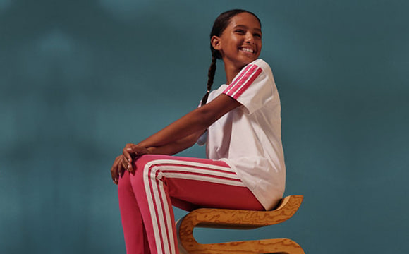 A young woman with braided hair wearing a white shirt and red and white striped pants stands against a teal background.