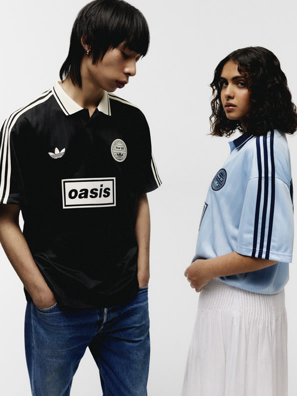 Two individuals, a young man and a young woman, are standing together in what appears to be a studio or photographic setting. The man is wearing a black Oasis-branded jersey, while the woman is wearing a light blue jersey with stripes.