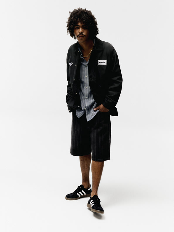 The image shows a person wearing a black jacket, a striped shirt, and black shorts, standing against a plain white background.