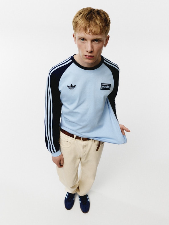 A young boy wearing a light blue Adidas shirt and khaki pants stands against a plain white background.