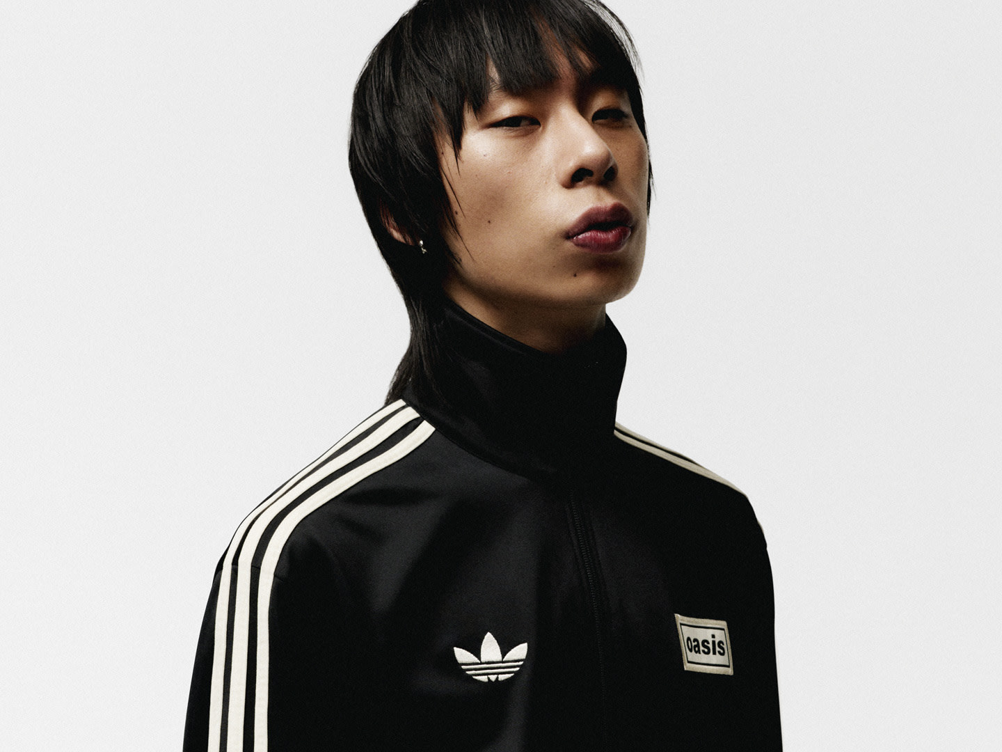 A young person with short, dark hair wearing a black Adidas jacket stands against a plain white background, their expression serious and focused.