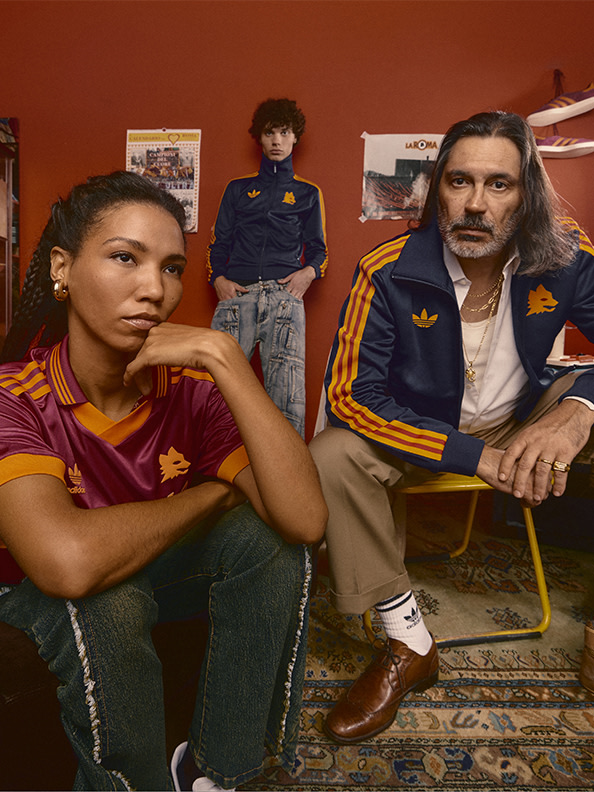 The image shows a group of people, including a woman with curly hair sitting in the foreground, a man with long hair sitting in the background, and a child standing in the middle. They are all wearing casual clothing and appear to be in a room with various items and decorations on the walls.