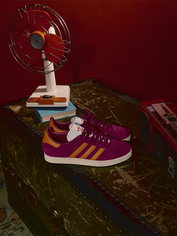 A pair of purple and orange Adidas sneakers sits on a wooden surface, accompanied by a vintage-style fan and various other objects in the background.