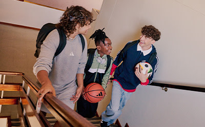 Three young individuals, two males and one female, are standing on a staircase holding various sports equipment, including a basketball.