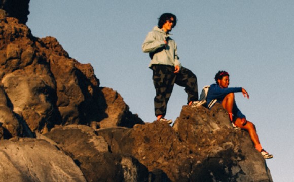 Two individuals standing on a rocky outcrop against a mountainous backdrop, with one person in a light-colored shirt and the other in a dark-colored shirt.