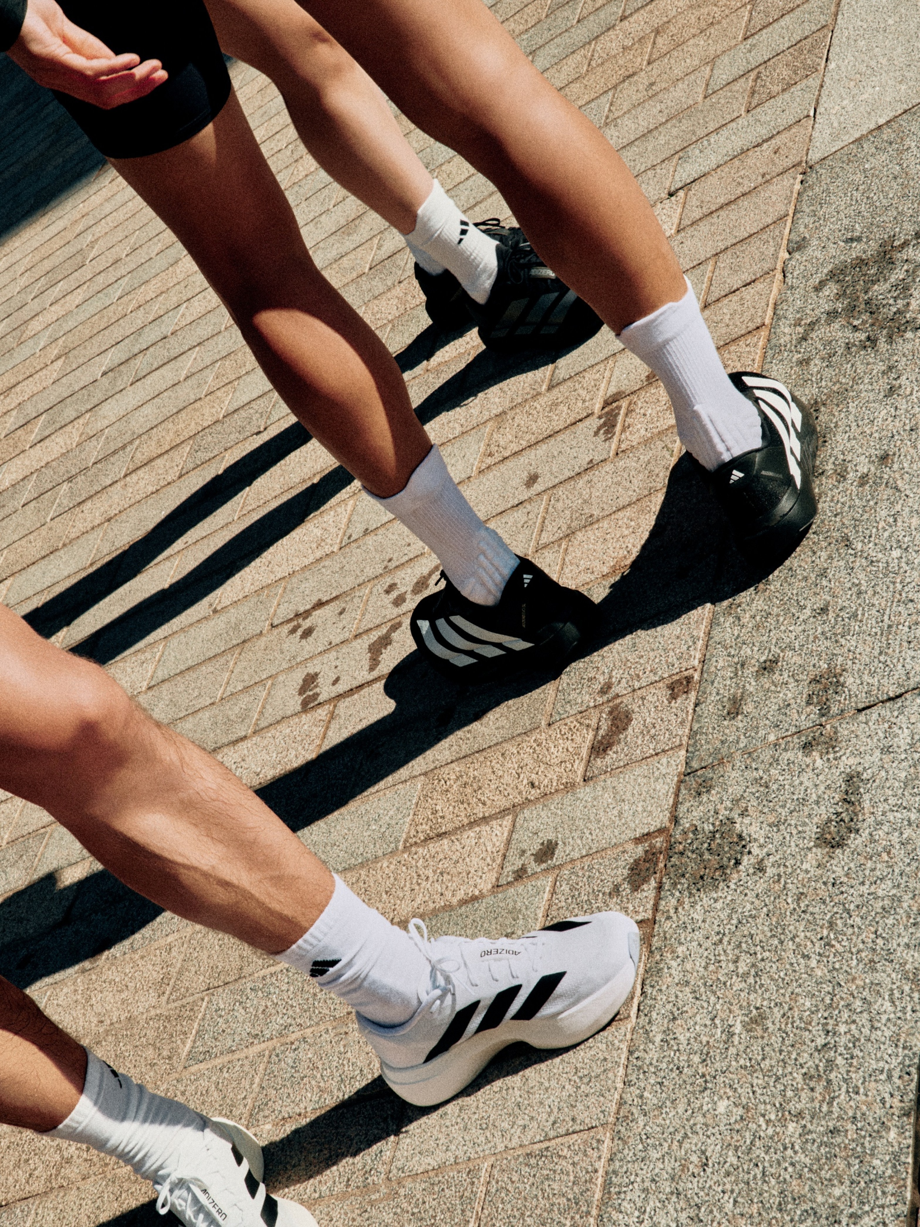 The image shows the lower body and feet of two people wearing athletic shoes and socks, standing on a brick surface.