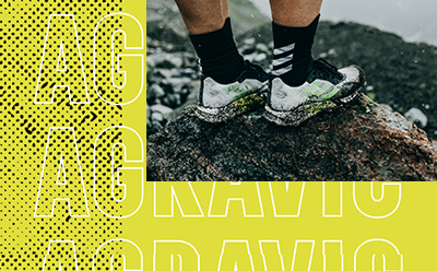 Die Füße einer Person mit schwarz-weißen Sneakern stehen auf einer felsigen, moosbewachsenen Oberfläche vor einem verschwommenen gelb-schwarzen Hintergrund.