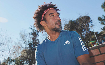 A man with curly hair wearing a blue Adidas shirt stands in front of a background of trees and a clear sky.