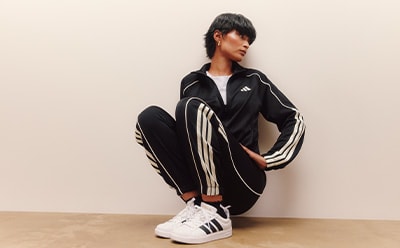 A young person wearing a black and white Adidas tracksuit jacket and pants, standing against a plain beige wall.
