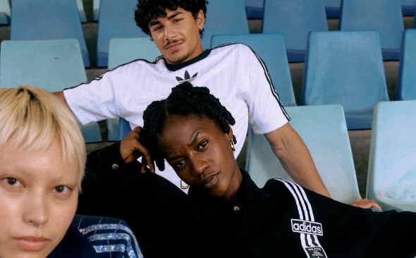 Three people standing in front of a large, printed canvas depicting an underground subway station. They all wear adidas Originals clothing and face different directions.