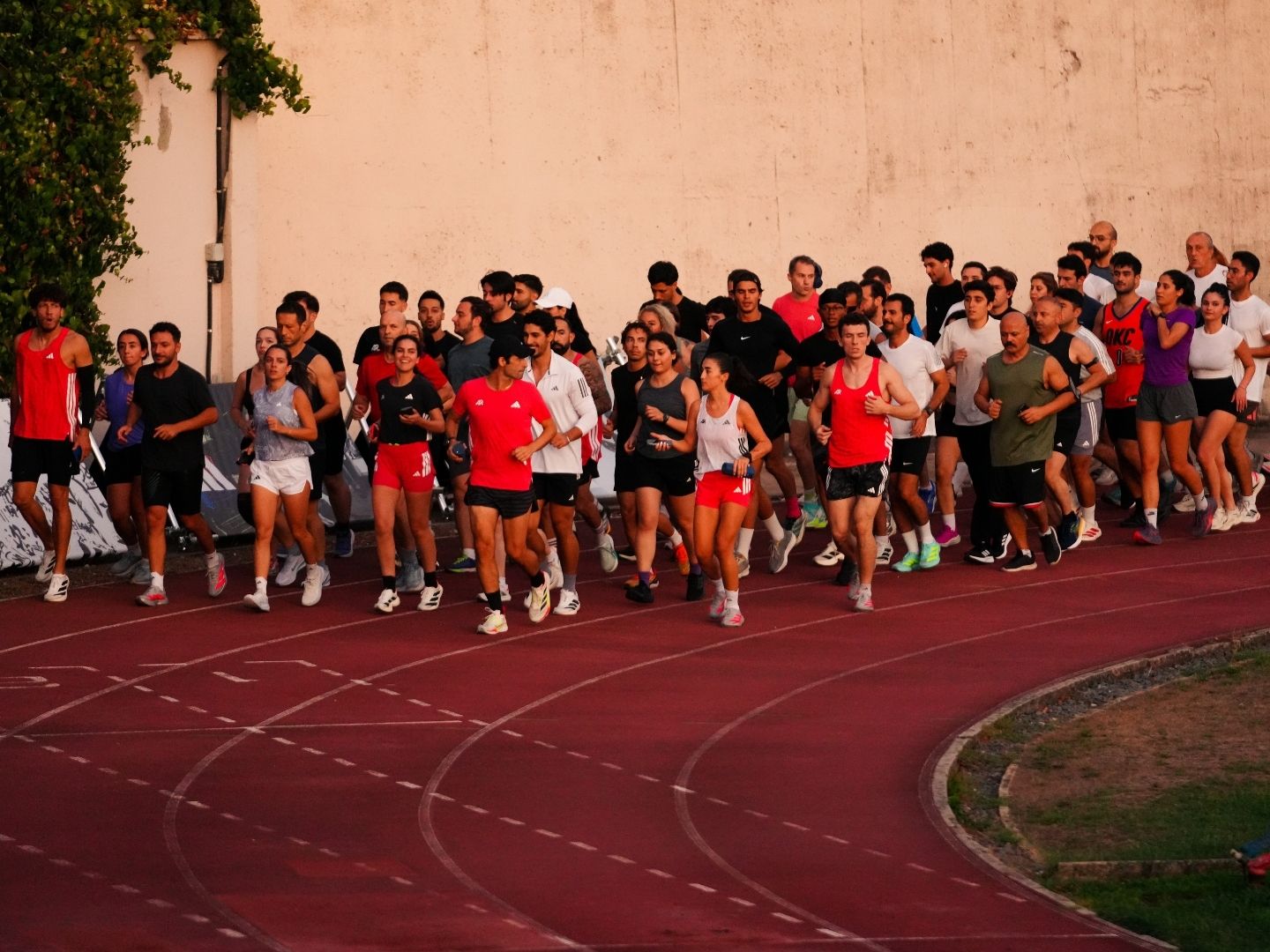 A large group of people, dressed in athletic attire, gathered on a red running track surrounded by greenery and a concrete wall.
