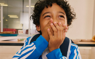 Un niño con el pelo rizado y una camisa a rayas azules y blancas que cubren su boca con las manos, parado en lo que parece ser un aula o un decorado educativo con estanterías y otros objetos visibles al fondo.