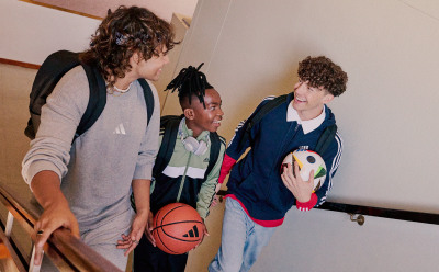 Drei junge Leute, zwei Jungen und ein Mädchen, stehen zusammen in einem Raum, der wie ein Flur oder eine Halle wirkt. Einer der Jungs hält einen Basketball in der Hand, was darauf hindeutet, dass sie vielleicht schon gespielt haben oder kurz davor stehen, ein Spiel zu spielen.