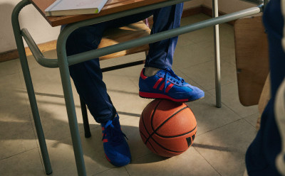 Debajo de un escritorio, en un aula, se ven un balón de baloncesto y un par de zapatillas.