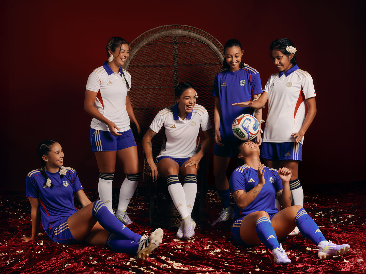 A group of female soccer players in uniform, posing together on a field covered in red leaves, with a large arched structure in the background.