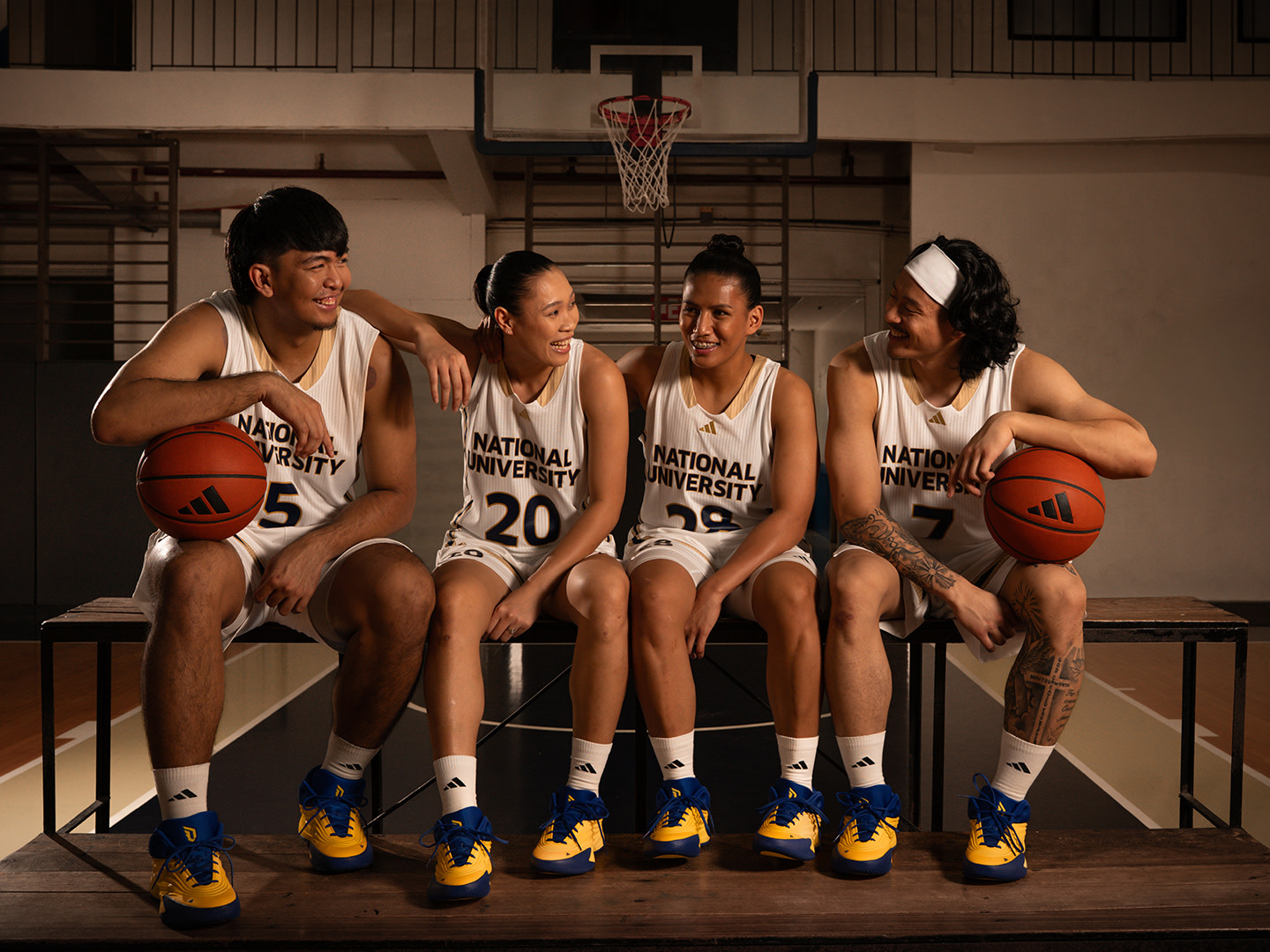 Four individuals, dressed in basketball uniforms, are seated on a bench in a gymnasium setting. The background includes a basketball hoop and other sports equipment, suggesting this is a basketball court or training facility.