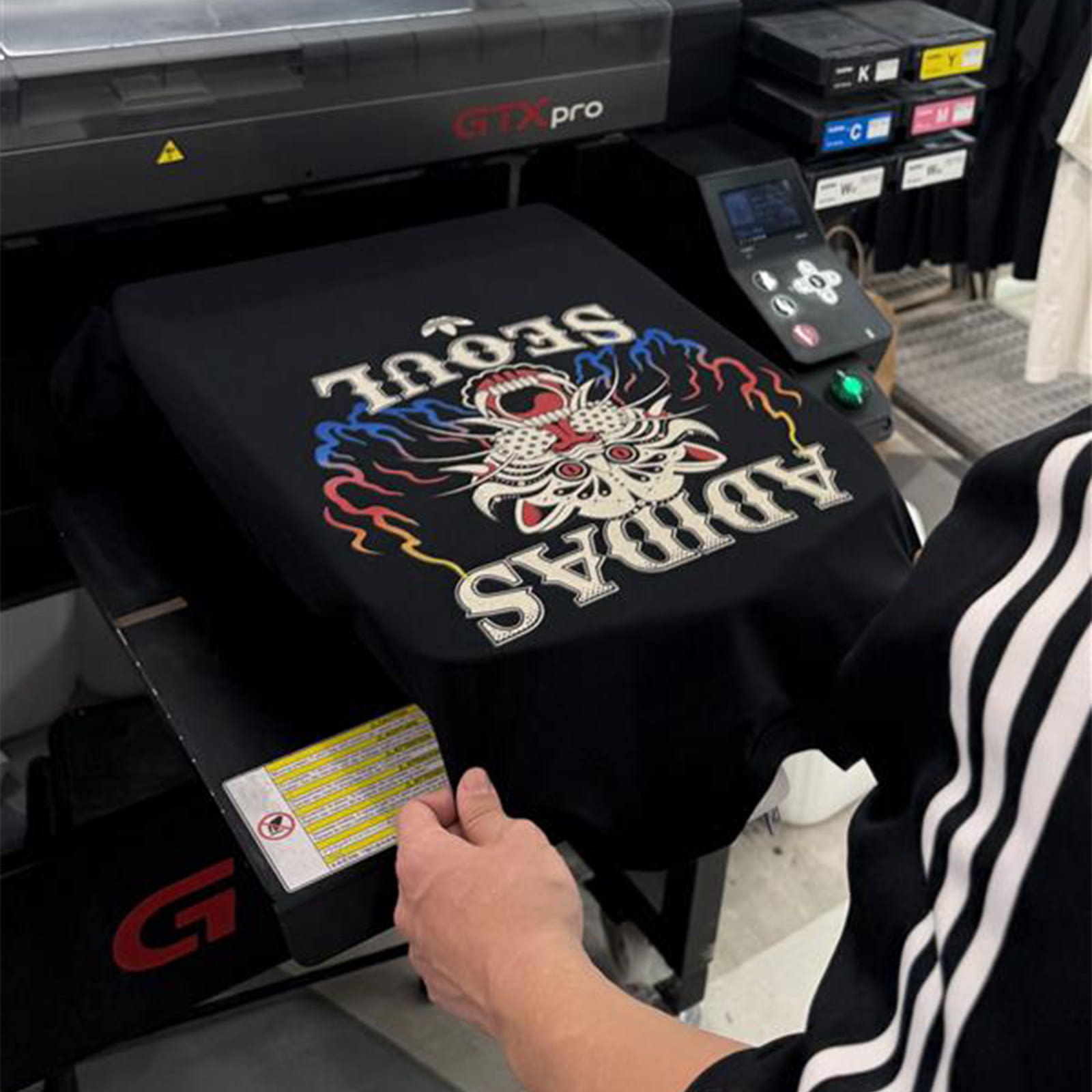 The image shows a black t-shirt with a colorful, intricate design of a skull and crossbones being printed on a large industrial printer.