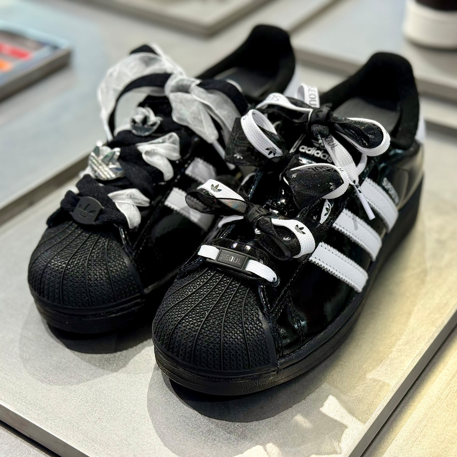 The image shows a pair of black and white Adidas Superstar sneakers with laces tied in a bow pattern, resting on a reflective surface.